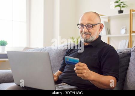 Smiling senior man pays online with credit card while using laptop at home Stock Photo