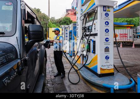 A petrol station attendant in a blue uniform refuels a car in New Delhi, with a red building and trees visible in the distance. Stock Photo