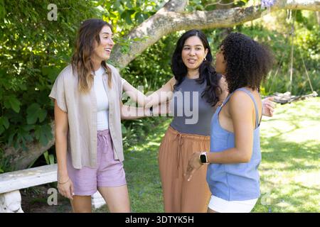 Diverse women friends talking in garden under tree near stone bench, wearing watches Stock Photo