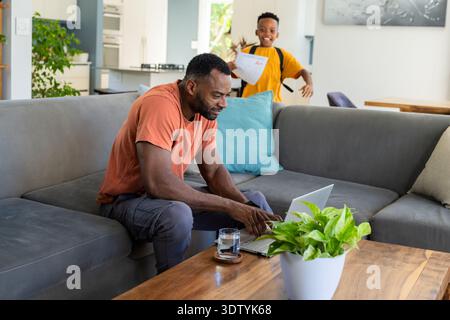 African American father typing on laptop at coffee table, schoolboy holding graded paper, backpack Stock Photo
