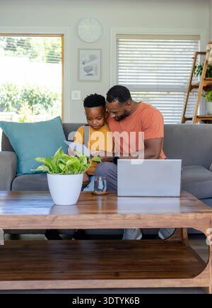 African American father and son reviewing school worksheet on gray sofa at home with open laptop Stock Photo
