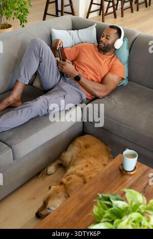 Mature African American man lying on sofa at home wearing orange-tee and white headset using phone Stock Photo
