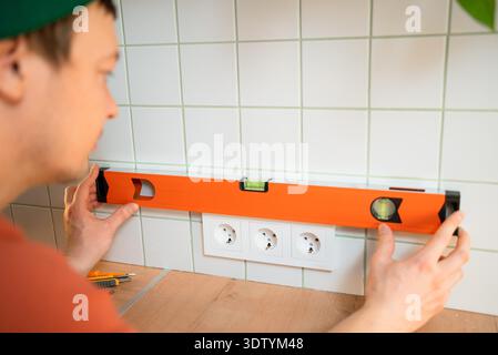 Electrician installing new power sockets into a tiled kitchen wall, carefully using a spirit level to ensure perfect horizontal alignment and professional quality for the home improvement project || Model released Stock Photo
