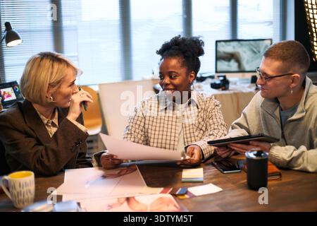 Middle aged Caucasian woman, young adult Black woman, young adult Caucasian man sitting at table discussing product photos during creative team meeting in modern office Stock Photo