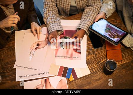Three young adult women and man, including Black and Caucasian, collaborating at table, discussing skincare product samples and color swatches, using digital tablet and printed materials Stock Photo