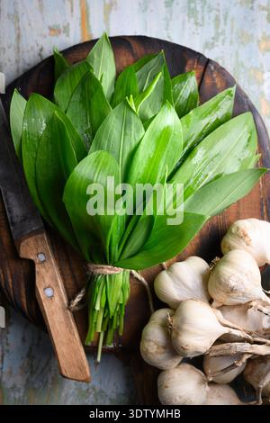 Fresh wild garlic leaves arranged on cutting board with kitchen knife and garlic bulbs. Raw garlic greens shown in food preparation scene. Flat lay, top view Stock Photo