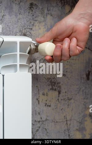 A human hand turns a cream-colored thermostat knob on a sleek white radiator, adjusting the temperature against a textured wall. Stock Photo