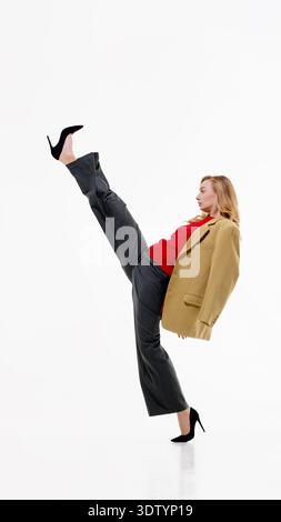 Woman in beige blazer, red sweater and gray trousers performing high kick on white background. Stock Photo