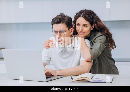 Couple working together on a laptop at home, showing teamwork and support, with a notebook nearby for notes. Woman provides emotional support to man who is tired and stressed from work. || Model released Stock Photo