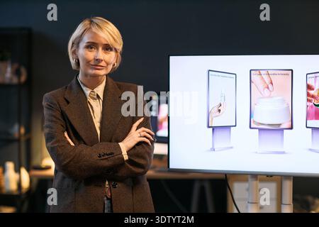 Caucasian middle aged woman standing with arms crossed in front of digital display showing skincare product advertisements, looking confidently at camera in modern workspace Stock Photo
