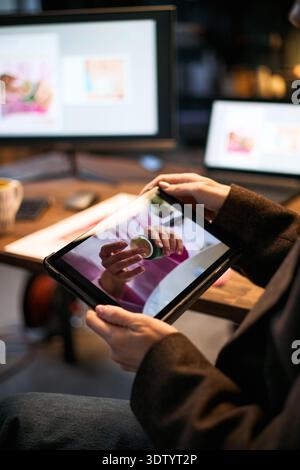 Caucasian woman holding digital tablet displaying close up of hands holding skincare product, sitting at desk with computer monitors in background, working in modern office Stock Photo