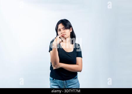 Portrait of a young Asian woman looking unsure or hesitant with hand on face. Concept of doubt, uncertainty, and decision making in studio setting. Stock Photo