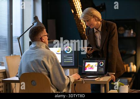 Caucasian young adult man sitting at desk working on graphic design project on laptop, Caucasian middle aged woman standing beside him holding digital tablet discussing work Stock Photo