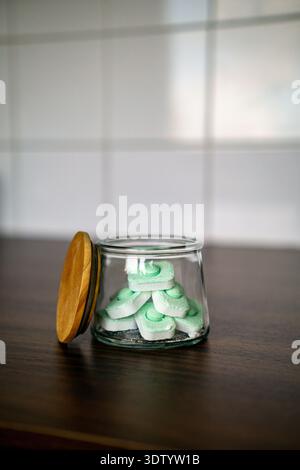 Transparent glass jar filled with green dishwasher tablets placed on a wooden surface. The wooden lid is open and resting beside the container. Stock Photo