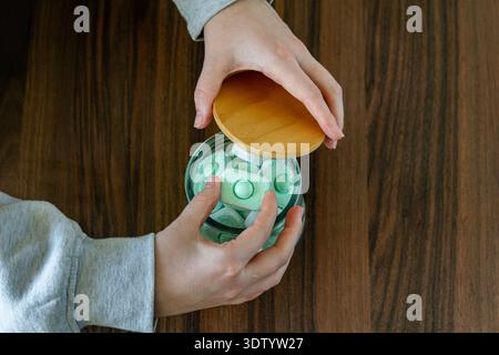 Top view of hands opening a glass jar filled with green dishwasher tablets on a wooden surface. Stock Photo