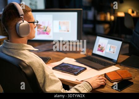 Caucasian young adult man wearing headphones working at desk using desktop computer and laptop with digital tablet and smartphone nearby focusing on creative project Stock Photo