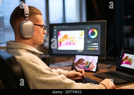 Caucasian young adult man wearing headphones using digital pen tablet while editing photo on computer monitor and tablet screen in modern workspace, focusing on creative work Stock Photo