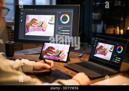 Caucasian young adult man using graphic tablet editing photo of Caucasian woman holding cosmetic jar on computer monitor, laptop, and tablet in modern workspace Stock Photo