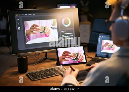 Caucasian young adult man using graphic tablet and stylus editing product photo on computer and tablet, wearing headphones, focusing on digital retouching process at desk Stock Photo