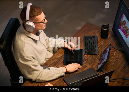 Caucasian young adult man wearing headphones using digital pen tablet while working on computer at desk, concentrating on creative project with stylus and multiple screens Stock Photo