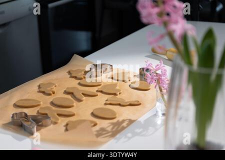 Unbaked gingerbread cookies in Easter bunny and egg shapes rest on baking paper. Metal cookie cutters and a rolling pin sit nearby on the bright white kitchen counter. Stock Photo