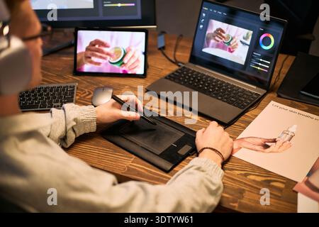 Caucasian young adult man using graphic tablet and stylus while editing product photo on laptop, tablet displaying same image, working at wooden desk with printed skincare advertisement Stock Photo