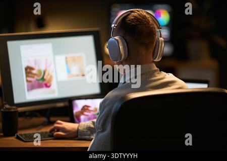 Caucasian young adult man wearing headphones sitting at desk working on computer with large monitor displaying images, focused on digital project in modern workspace Stock Photo