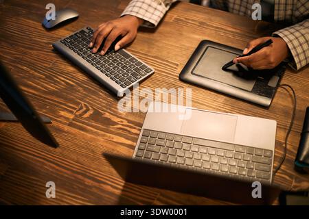 Black woman using digital drawing tablet and wireless keyboard while working at desk, hands visible interacting with technology, likely engaged in creative or design task, overhead view Stock Photo