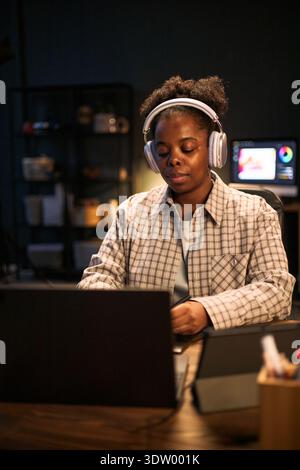 Young adult Black woman wearing headphones working on laptop at desk, concentrating on screen with digital tablet and computer monitor in background, hands on keyboard Stock Photo