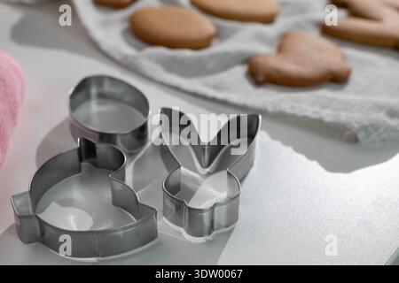 Some cookie cutters shaped like bunnies sit on a light table. In the background, baked gingerbread cookies lay on a towel. The scene shows Easter baking during a celebration. Stock Photo