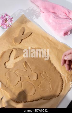 A person bakes festive Easter gingerbread cookies, cutting bunny and egg shapes from light brown dough on parchment paper. Stock Photo