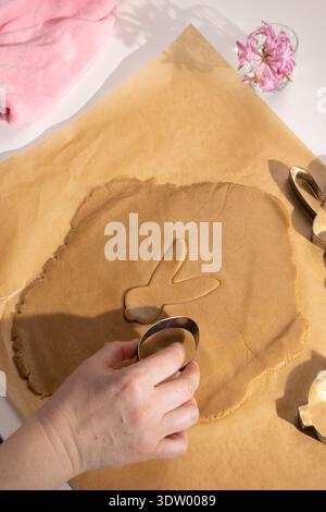 A person's hand presses a round cutter into brown gingerbread dough on parchment paper, next to an already cut Easter bunny shape. Stock Photo