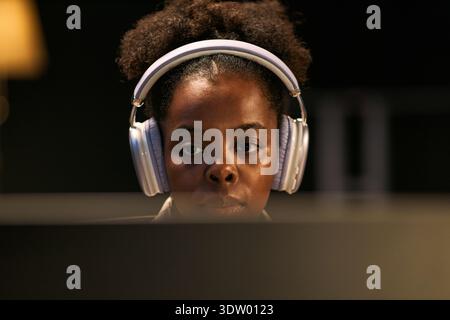 Portrait of young adult Black woman wearing headphones concentrating while looking at computer screen in dark environment, focused expression visible on face Stock Photo