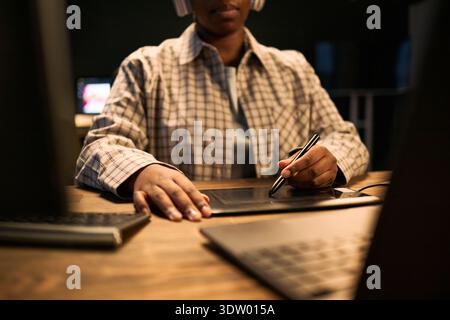 Black young adult woman using digital pen on graphic tablet while working at desk with laptop and monitor, wearing headphones, focusing on creative project in modern workspace Stock Photo