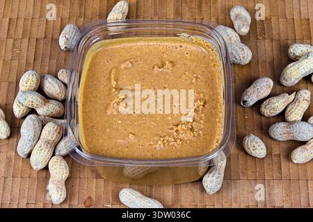 A close-up overhead view of creamy peanut butter in a plastic tub with whole peanuts scattered around it on a wooden surface. Stock Photo