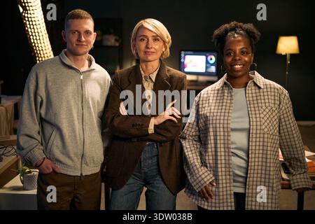 Portrait of young Caucasian man, middle aged Caucasian woman and young Black woman standing together smiling at camera in creative office environment with computer screen in background Stock Photo