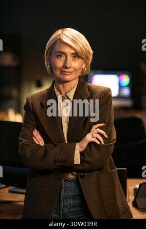 Portrait of middle aged Caucasian woman standing with arms crossed, smiling confidently while looking into camera, office environment with computer monitor in background visible Stock Photo