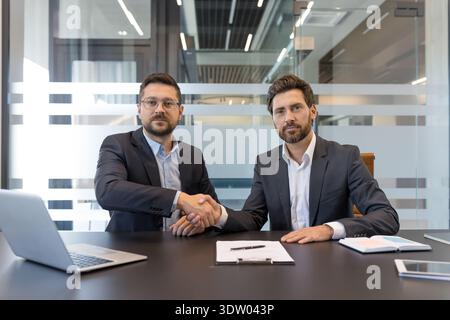 Two confident businessmen shaking hands over a desk with a laptop and documents, symbolizing a successful partnership agreement or the completion of a professional deal in a modern office Stock Photo