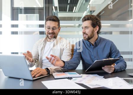 Two businessmen discussing project details and ideas while working together on a laptop, fostering teamwork and professional collaboration in a modern office environment Stock Photo