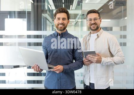 Two smiling male business colleagues standing in a modern office, holding a laptop and a digital tablet, reflecting teamwork and success in a professional environment Stock Photo