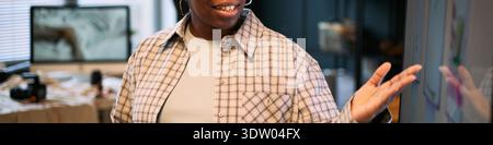Young adult Black woman smiling while interacting with touchscreen display in modern office setting, gesturing with right hand, visible technology and blurred workspace in background Stock Photo