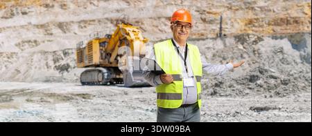 Smiling mining engineer in safety gear presenting a large excavator in a quarry Stock Photo