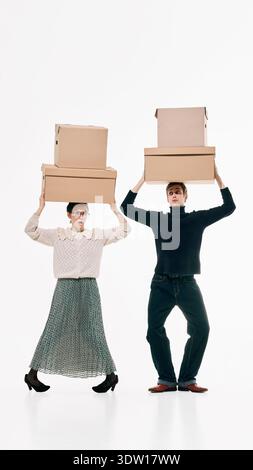 Stylish man and woman balancing stacked cardboard boxes on heads against white background. Stock Photo