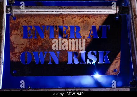 Rusty metal warning sign reading "enter at own risk" with blue lighting and industrial background, conveying danger and restricted access. Stock Photo