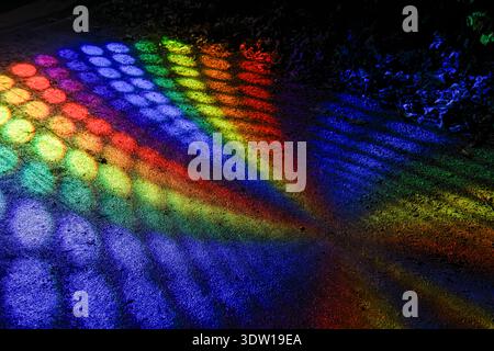 Rainbow spectrum light pattern with circular shadows cast on rough pavement at night, abstract colorful projection creating vivid stripes and dots. Stock Photo
