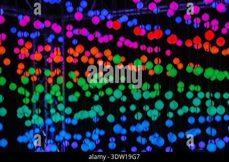 Abstract display of hanging multicolored glowing spheres on strings against dark background, creating vibrant bokeh pattern, modern light installation Stock Photo
