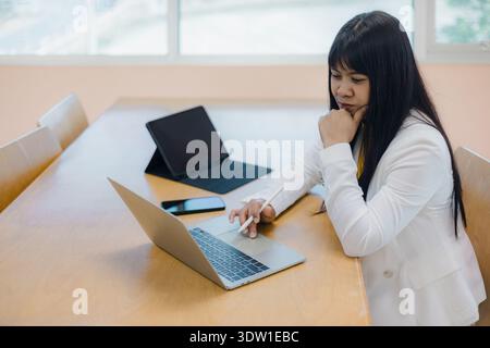 Asian businesswoman analyzing data on laptop and tablet in office. Professional female using smartphone and stylus pen for digital tasks at workplace. Stock Photo