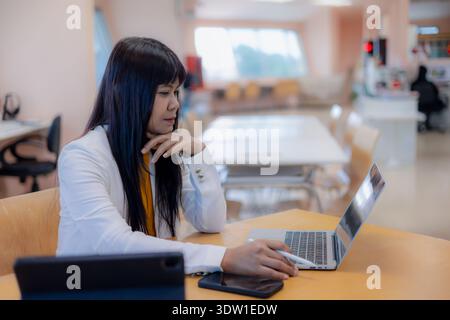 Pensive Asian businesswoman looking at laptop screen and thinking in office. Concentrated female professional working on project using digital technol Stock Photo