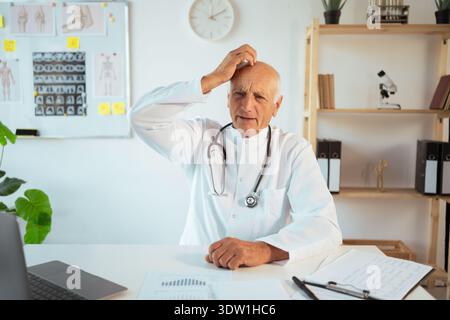 Stressed Senior man doctor scratch head making mistake working in clinic on laptop computer. healthcare professional in office Stock Photo