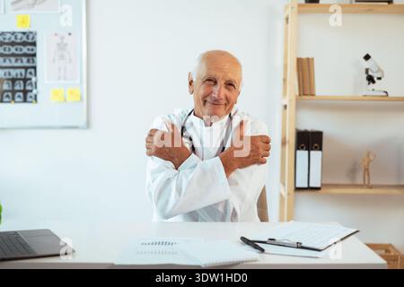 Senior man doctor leads healthy consultation in clinic hugging himself and smiling, feeling comfortable discussing with patient looking at camera Stock Photo